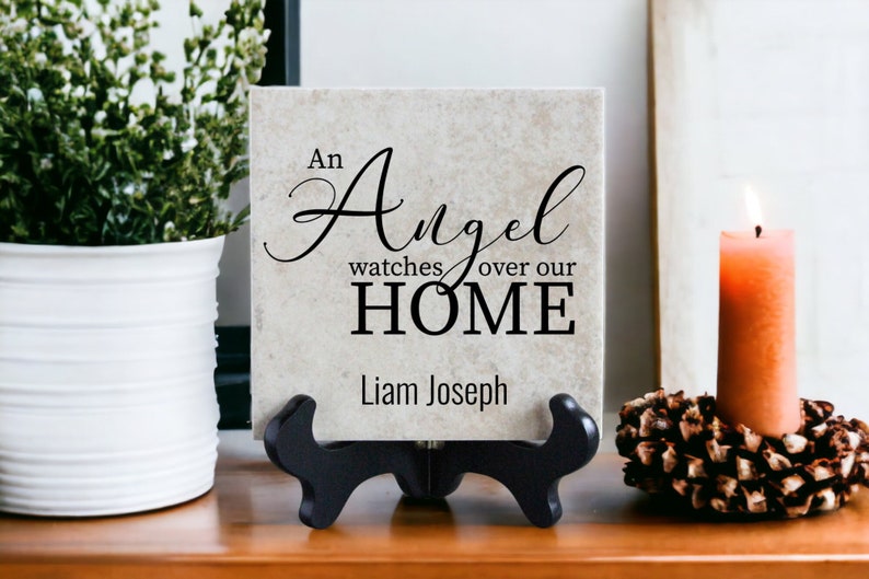 May include: A beige ceramic tile with black text that reads "An Angel watches over our HOME" and "Liam Joseph". The tile is on a black stand and there is a lit candle and a pine cone decoration on a wooden surface.