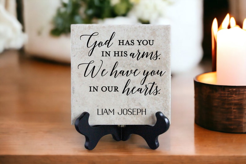 May include: A beige ceramic tile with black text that reads "God has you in his arms. We have you in our hearts. Liam Joseph". The tile is on a black stand and a wooden surface.