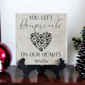 May include: A beige ceramic tile with black text that reads "You left pawprints on our hearts" and the name "Wolfie". The tile is on a black stand and has a heart shape made of paw prints.