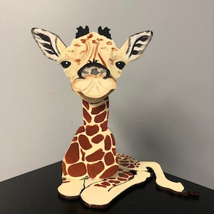 May include: A wooden giraffe figurine painted with brown and white spots. The giraffe has a long neck and is sitting down with its legs tucked under its body. The giraffe has a cute, cartoon-like expression.