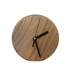 May include: A circular wooden clock with a light brown finish. It features black hands and a silver centre. The clock face is made of wood, with visible grain. The design is simple and modern. The clock's diameter is approximately 15 cm.