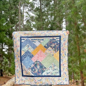 May include: A colourful quilt featuring a mountain design. The quilt is made from a variety of floral fabrics in shades of blue, green, pink, yellow and white. The quilt is framed with a white border and a navy blue inner border.
