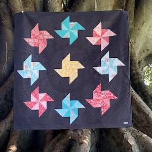 Op de afbeelding: Een zwarte quilt met een cirkelvormig patroon van negen kleurrijke windmolens. Elke windmolen is gemaakt van stof met een bloemenprint en heeft een ander kleurenschema. De quilt hangt aan een boomstam.