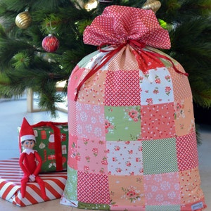 May include: A large red and pink patchwork Christmas gift bag with a drawstring closure. The bag is made of various fabrics with floral and geometric patterns. The bag is sitting on a wooden floor with a Christmas elf toy in the foreground.