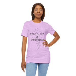 May include: A lilac t-shirt featuring the text "REGULATED & UNBOTHERED" in black, accompanied by a heart, crown, and lightning bolt graphic. The tee is made of a soft fabric, suitable for everyday wear.