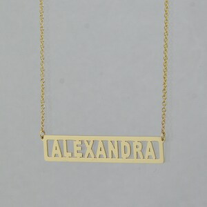 May include: A gold necklace with a rectangular pendant that says "ALEXANDRA" in cutout letters.