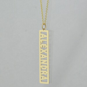 May include: A gold necklace with a long, rectangular pendant that has the name "ALEXANDRA" cut out of it.