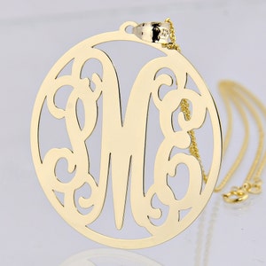 May include: A gold-colored pendant necklace featuring a large, circular design with an ornate letter "M" in the center. The pendant hangs from a delicate gold chain. The design includes decorative swirls and cut-out details.