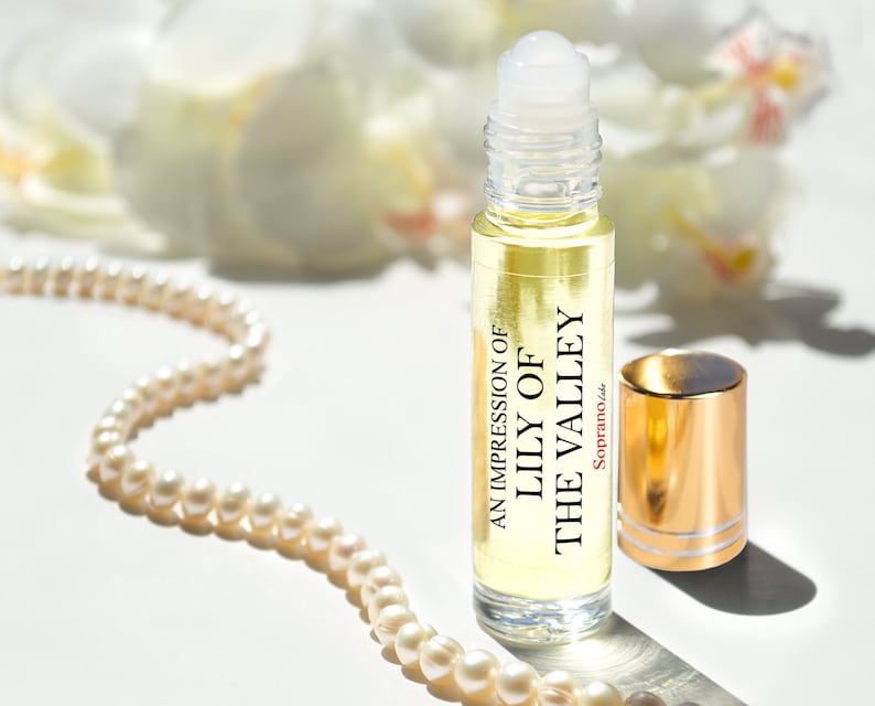May include: A clear glass roller bottle with a gold cap and a white label that reads "An Impression of Lily of the Valley" and "Soprano, Inc.". The bottle is sitting on a white surface with a pearl necklace.