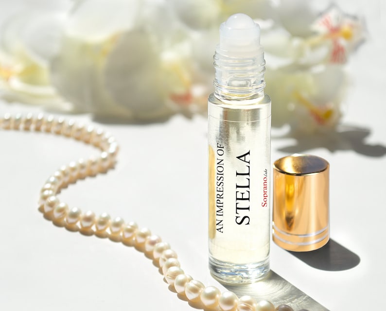 Może przedstawiać: Przezroczysta szklana butelka z rolką z złotą zakrętką i białą etykietą z napisem "An Impression of Stella Soprano, Inc.". Butelka stoi na białym tle z naszyjnikiem z pereł.