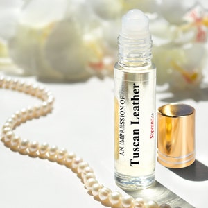May include: A clear glass roller bottle with a gold cap and a white label that reads "An Impression of Tuscan Leather Soprano, Ltd."
