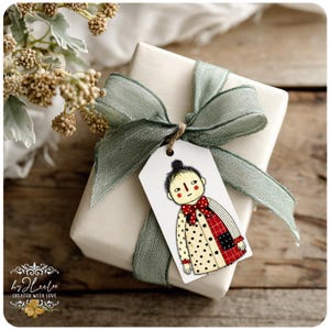 Puede incluir: Un regalo envuelto con una cinta verde salvia y una etiqueta con una ilustraci&oacute;n caprichosa de una persona con un abrigo de lunares y una pajarita roja. La etiqueta est&aacute; atada con hilo. El regalo est&aacute; sobre una superficie de madera.