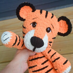 May include: A plush tiger toy made of soft orange yarn with black stripes and a white muzzle. The stuffed animal features large dark brown plastic eyes, rounded ears with black edges, and a black yarn nose. One arm is raised in a waving gesture.