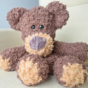 May include: A plush teddy bear crafted from textured dark brown yarn with a lighter tan muzzle and matching tan accents on the paws. The bear features two dark, round plastic eyes and a prominent, vertically ribbed purple nose centered on the face.