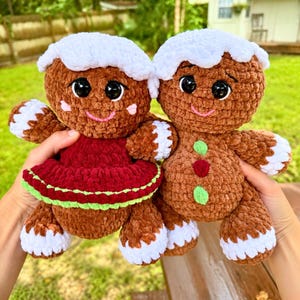 May include: Two crocheted gingerbread dolls made of brown yarn with white frosting-like accents on the head and limbs. One doll wears a dark red dress with a green trim, while the other features three small green and red circular buttons.