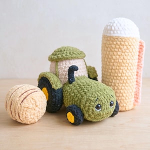 Peut inclure: Ensemble de jouets crochetés faits à la main. On distingue un tracteur vert avec des roues noires et jaunes, un silo beige et vert, une balle beige à rayures marron et une grande chope de bière avec une anse rose.