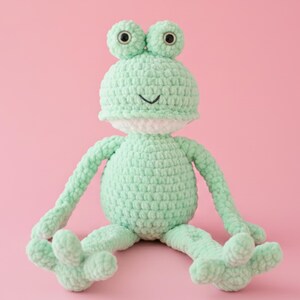 Puede incluir: Un peluche de rana tejido a ganchillo en color verde menta claro con cabeza redonda, grandes ojos de plástico negro y una mancha blanca en el vientre. El juguete tiene brazos y piernas largos y delgados con pies de tres dedos.