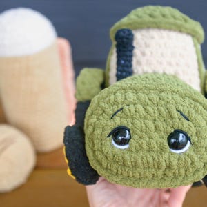 May include: A crochet tractor toy made with olive green yarn, featuring a cream-colored cab, a black chimney stack, and black wheels. The front of the tractor has large, glossy black safety eyes with white accents and embroidered eyebrows.