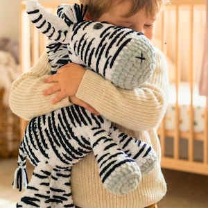 May include: A plush zebra stuffed animal in white with black stripes is being held. The zebra has a gray muzzle and feet. The stuffed animal is being held by a person wearing a cream-colored sweater. A wooden crib is in the background.