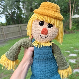 May include: A handmade crochet scarecrow doll featuring a yellow hat, bright yellow yarn hair, and a tan face with a stitched orange nose. The doll wears dark teal overalls over olive green sleeves, with yellow fringe at the wrists and neck.