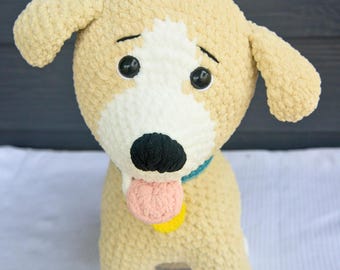 May include: A plush crochet dog toy made of tan and white yarn with a soft, fuzzy texture. The dog features large black plastic eyes, a black stitched nose, a small pink tongue, a teal collar, and a round yellow tag hanging from the collar.