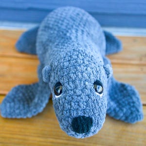 May include: A handmade, gray crocheted seal plush toy with black eyes and a black nose. The seal is lying down on a wooden surface, with its flippers extended. The plush toy is soft and cuddly, perfect for children.
