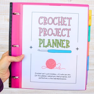 May include: A pink binder with a black spine holds a white page titled "CROCHET PROJECT PLANNER." The page features a crochet hook, a ball of yarn, and text that reads "Crochet isn't just a hobby—it's who you are."
