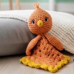May include: A handmade crochet chick toy in shades of orange and yellow. The chick has a round head with black eyes and a yellow beak. The body is a blanket-like shape with a scalloped yellow border. The toy is made of soft yarn.