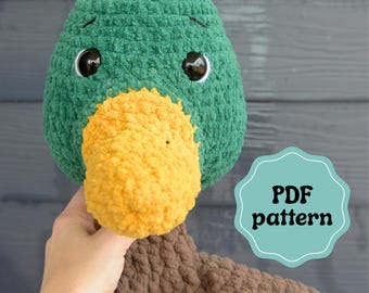 May include: A crocheted duck plush toy featuring a textured green head, a bright yellow beak, and a brown body. The toy has large black safety eyes and embroidered eyebrows. A light blue scalloped label displays the text PDF pattern.