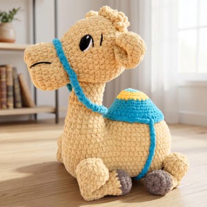 Puede incluir: Un camello de peluche hecho de hilo color canela con un acabado de ganchillo texturizado. Presenta una brida azul brillante, una silla de montar azul y amarilla en el lomo y detalles en gris oscuro en las patas. Tiene grandes ojos negros.