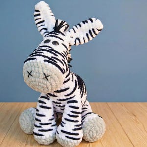 May include: A handmade, plush zebra toy with black and white stripes. The zebra has large, floppy ears, a gray muzzle with stitched "X" marks, and gray feet. The toy is sitting on a wooden surface against a blue background.