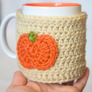 May include: A white ceramic mug with an orange interior, featuring a beige crocheted cozy sleeve. The sleeve is decorated with a textured, orange crocheted pumpkin applique that has a small green stem attached to the top center of the pumpkin.