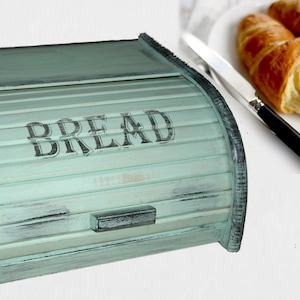 May include: A distressed, mint green bread bin with the word "BREAD" stencilled on the front. The bin has a roll-top design and a small handle. A plate with croissants and a knife are in the background.