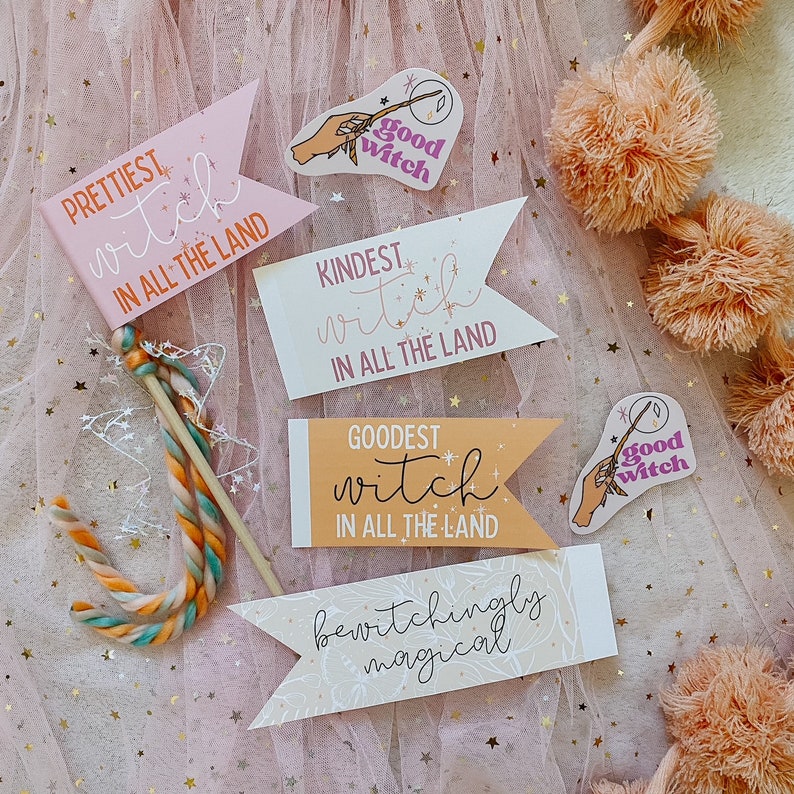 Puede incluir: Un conjunto de cinco banderas de papel rosa, naranja y blanco con purpurina dorada y estrellas. Las banderas tienen el texto "PRETTIEST witch IN ALL THE LAND", "KINDEST witch IN ALL THE LAND", "GOODEST witch IN ALL THE LAND" y "bewitchingly magical". Tambi&eacute;n hay dos pegatinas con el texto "good witch" y una mano sosteniendo una varita.