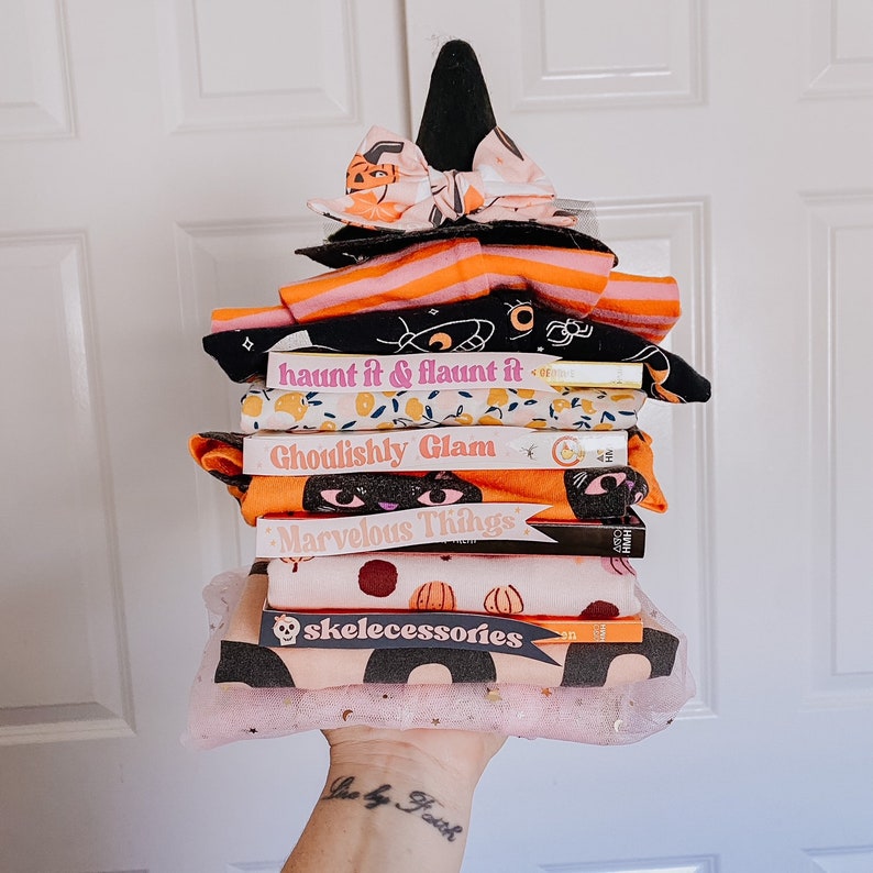 Puede incluir: Una pila de libros con portadas de tem&aacute;tica de Halloween, que incluyen "haunt it & flaunt it", "Ghoulishly Glam" y "Marvelous Things". Los libros est&aacute;n envueltos en una tela de tul rosa con un sombrero de bruja negro y un lazo naranja y negro en la parte superior.