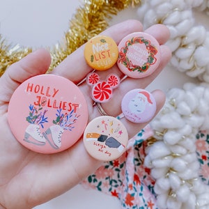 May include: A hand holding a collection of six Christmas themed button pins. The pins are pink, red, and white with various holiday designs, including a holly sprig, a snowman, a candy cane, and a wreath. The text on the pins reads "Holly Jolliest", "Merry and Bright", "It's a Jolly Holiday", "Sleigh All Day", and "Nice List Club".