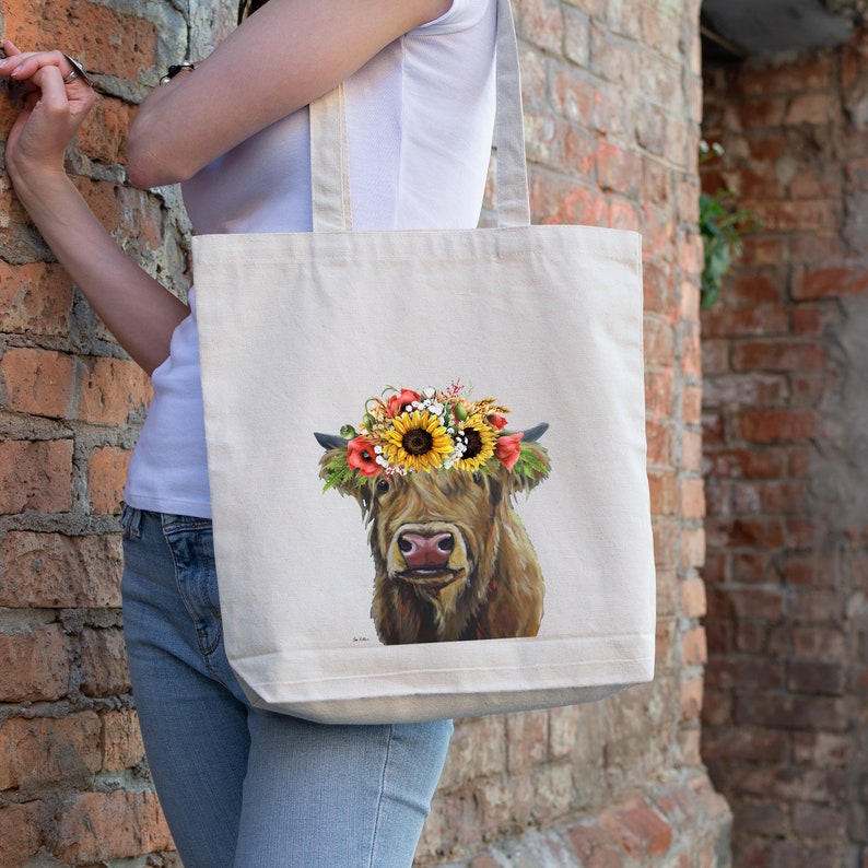 May include: A white canvas tote bag with a brown Highland cow wearing a flower crown. The crown is made of sunflowers, poppies, and other wildflowers.