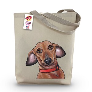 Pu&ograve; includere: Una borsa tote in tela beige con un'illustrazione di un bassotto rosso e marrone. La borsa ha un'etichetta che dice "Hippie Hound Studios" e "Made in the USA".