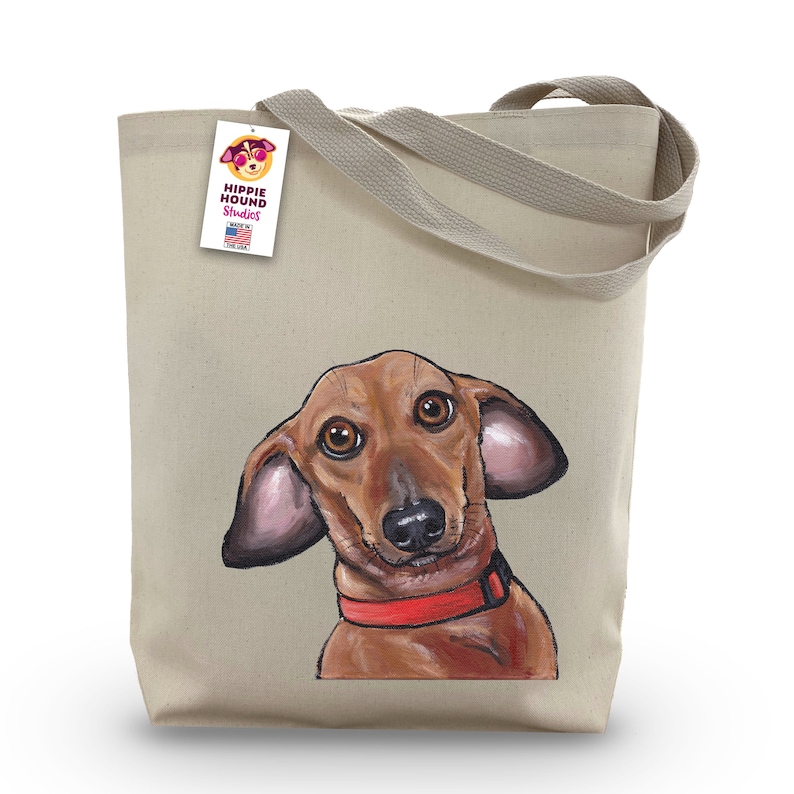 Pu&ograve; includere: Una borsa tote in tela beige con un'illustrazione di un bassotto rosso e marrone. La borsa ha un'etichetta che dice "Hippie Hound Studios" e "Made in the USA".