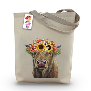 May include: A beige canvas tote bag with a floral crown on a brown Highland cow. The bag has a tag that says "Hippie Hound Studios" with a small American flag.