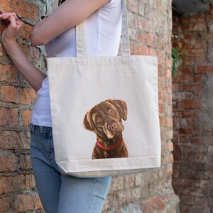 May include: A white canvas tote bag featuring a brown and white illustration of a chocolate Labrador Retriever wearing a red collar. The dog is looking up and to the right.