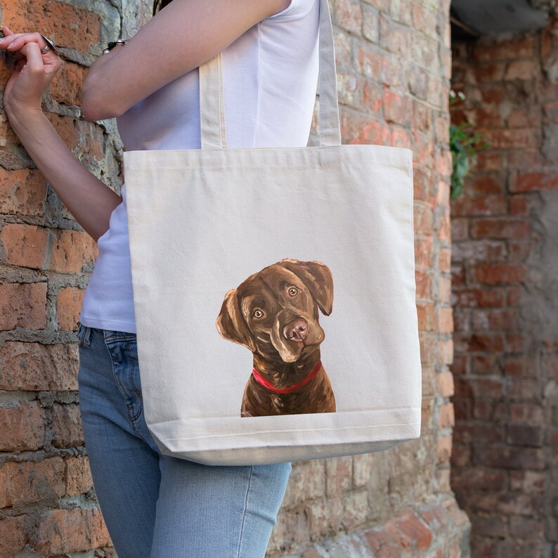 May include: A white canvas tote bag featuring a brown and white illustration of a chocolate Labrador Retriever wearing a red collar. The dog is looking up and to the right.