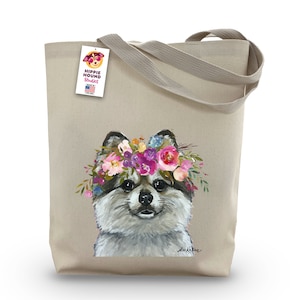 May include: A beige canvas tote bag with a floral crown on a fluffy white dog. The dog is looking at the viewer. The bag has a tag that says "Hippie Hound Studios" and "Made in the USA".