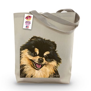 Può includere: Una borsa tote in tela beige con un'illustrazione di un cane pomerania nero e marrone. Il cane guarda lo spettatore con la lingua di fuori. La borsa ha un'etichetta che dice "Hippie Hound Studios" con una piccola bandiera americana.