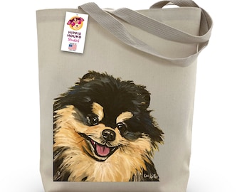 May include: A beige canvas tote bag featuring an illustration of a black and tan Pomeranian dog. The dog is looking at the viewer with its tongue out. The bag has a tag that reads "Hippie Hound Studios" with a small American flag.