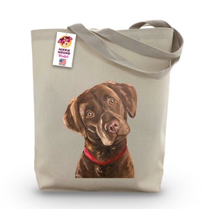 May include: A brown Labrador Retriever dog with a red collar is printed on a beige canvas tote bag. The bag has a tag attached to the handle that says "Hippie Hound Studios" and "Made in the USA".