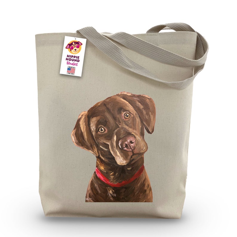 May include: A brown Labrador Retriever dog with a red collar is printed on a beige canvas tote bag. The bag has a tag attached to the handle that says "Hippie Hound Studios" and "Made in the USA".