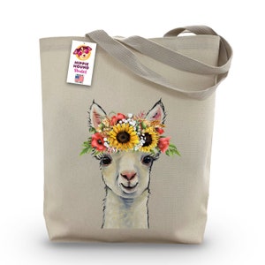 Può includere: Una borsa tote in tela beige con un lama bianco che indossa una corona di fiori. La corona è fatta di girasoli, papaveri rossi e fiori bianchi. La borsa ha un'etichetta che dice "Hippie Hound Studios" con una piccola bandiera americana.