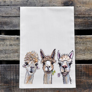May include: A white kitchen towel with a hand-painted illustration of three alpacas. The alpacas are brown and white with black eyes and noses. One alpaca is holding a small bouquet of yellow flowers in its mouth.