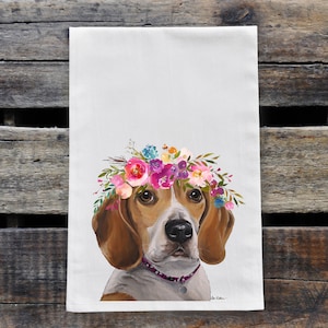 May include: White kitchen towel featuring a watercolor painting of a beagle wearing a floral crown. The dog has brown and white fur, with a pink collar. The floral crown includes pink, purple, blue, and yellow flowers. The towel is displayed on a wooden surface.